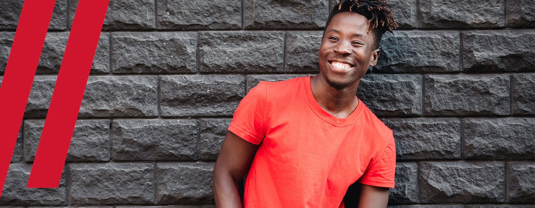 Photograph of a man wearing a red shirt and leaning against a black stone wall.  He has a big smile on his face. 